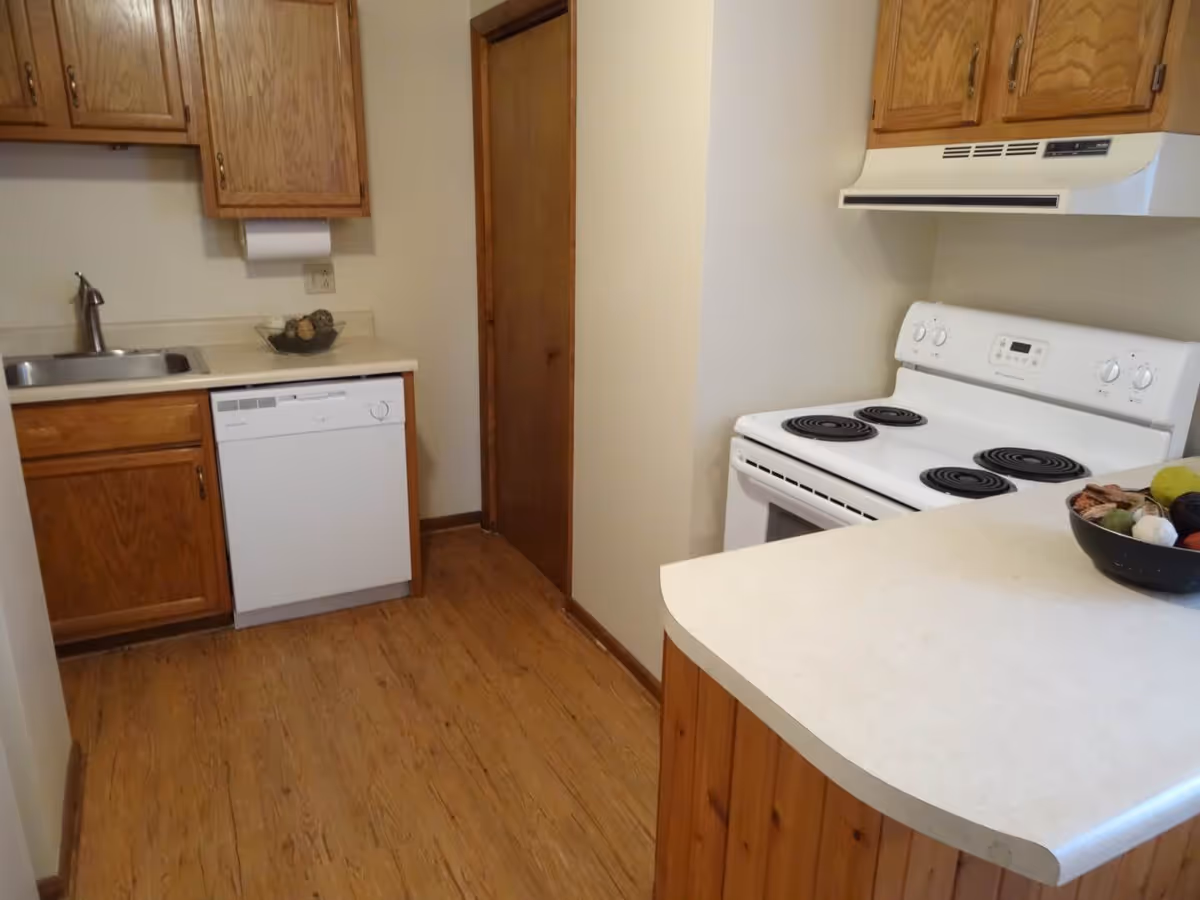 A small kitchen with wooden cabinets, a white electric stove with four coil burners, a white dishwasher, a stainless steel sink, and a countertop with a bowl of decorative items. The floor is wooden, and there is a wooden door in the background.