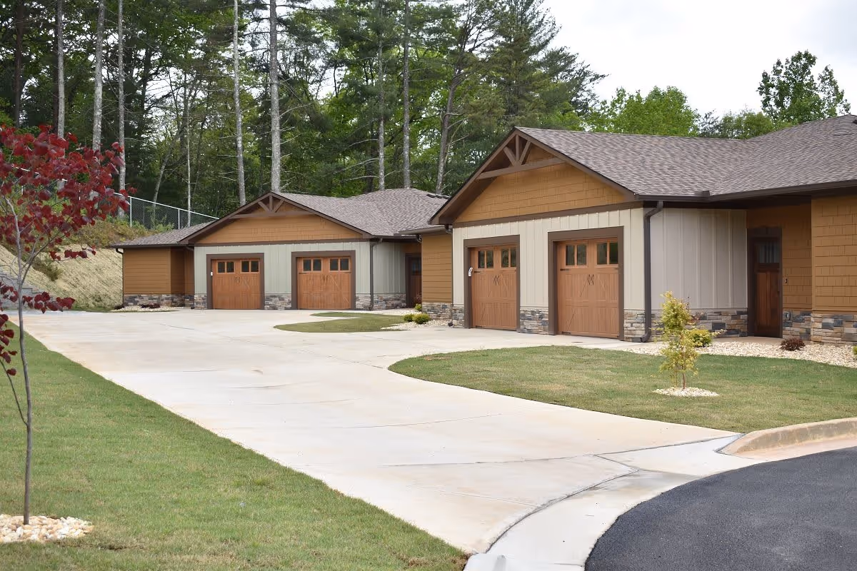 Exterior view of a senior living facility named Hiawassee Park showing a driveway leading to multiple garages with wooden doors. The building has a combination of tan and brown siding with stone accents at the base. There are small landscaped areas with grass, young trees, and shrubs, and tall trees in the background.