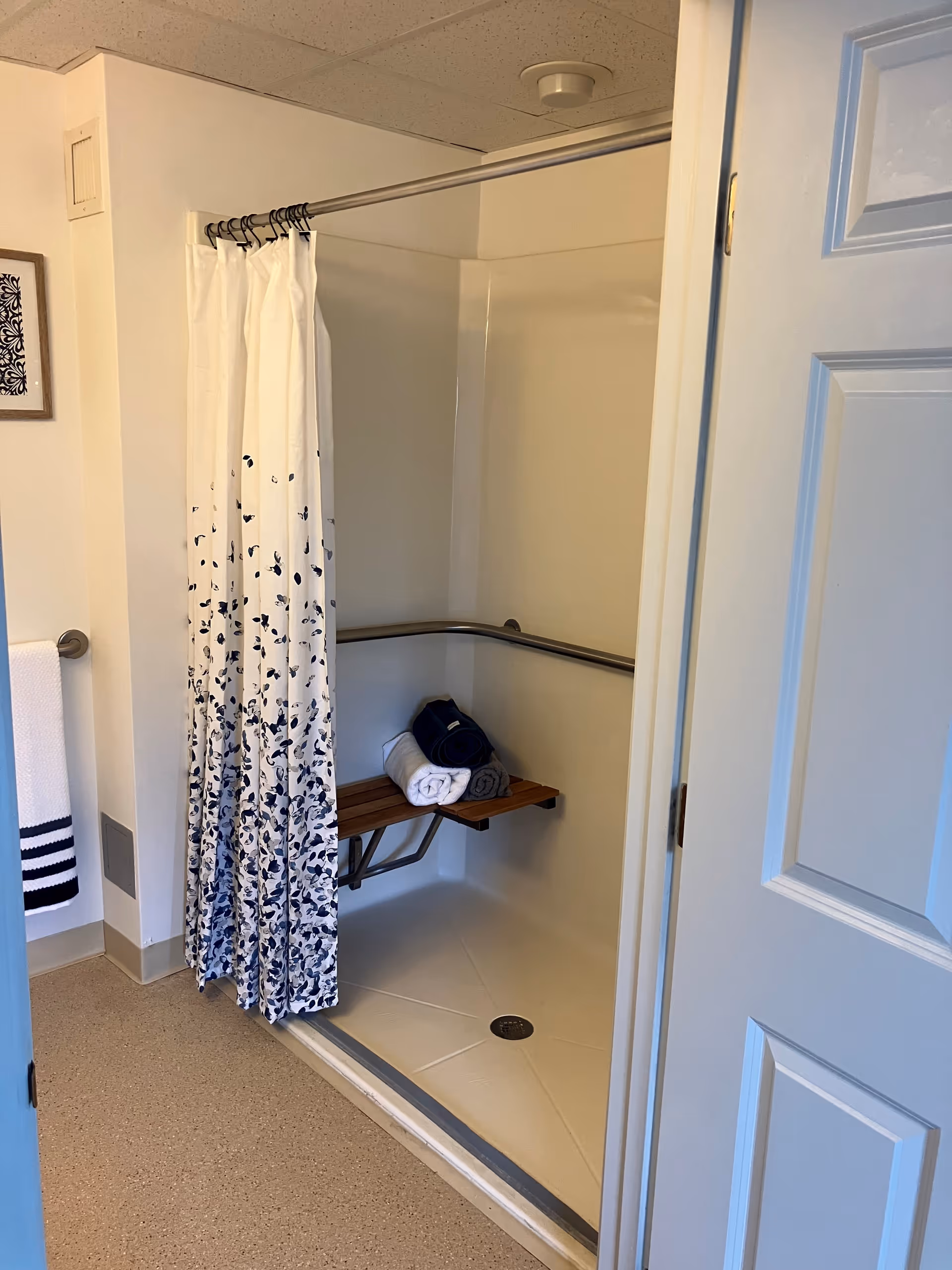 A bathroom shower area with a white and blue patterned shower curtain partially drawn to the side. Inside the shower, there is a wooden fold-down bench with three rolled towels in navy blue, white, and gray placed on it. The shower has a grab bar along the walls for support. The floor outside the shower is a speckled beige color, and a white door is partially open to the right.