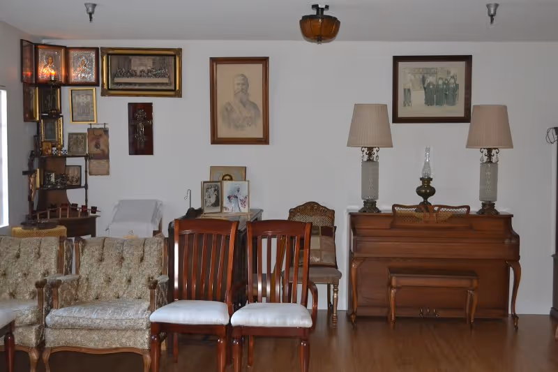 A room with vintage furniture including two upholstered armchairs, two wooden chairs with white cushions, a wooden piano with a matching bench, and two table lamps on top of the piano. The walls are decorated with framed pictures and portraits, and there is a small table with additional framed photos and a chair in the corner.