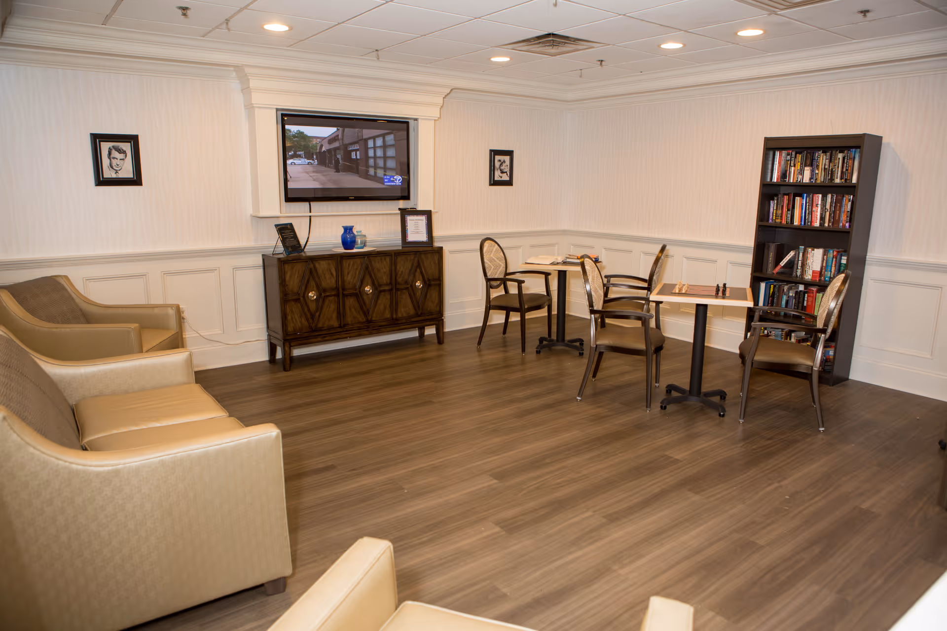 A cozy common area with beige armchairs and a sofa arranged around a wooden cabinet with a mounted flat-screen TV above it. Two small tables with chairs are set up near a bookshelf filled with books. The room has wood flooring, white paneled walls, and recessed ceiling lights.
