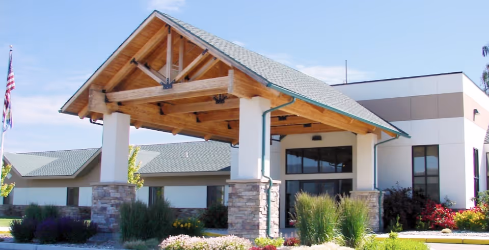 Exterior view of Clark Fork Valley Nursing Home featuring a covered entrance with wooden beams and stone pillars, surrounded by landscaped greenery and flowers under a clear blue sky.