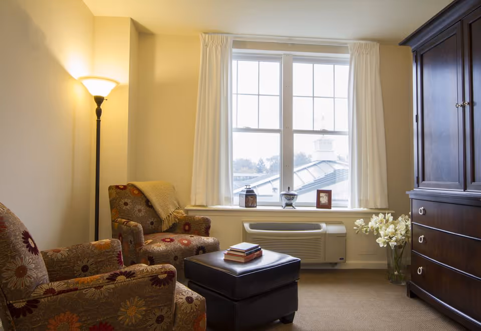 A cozy sitting area in a room with two floral-patterned armchairs, a black ottoman with books on top, a tall floor lamp, a large window with white curtains, a wooden cabinet, and a vase of white flowers on the floor.