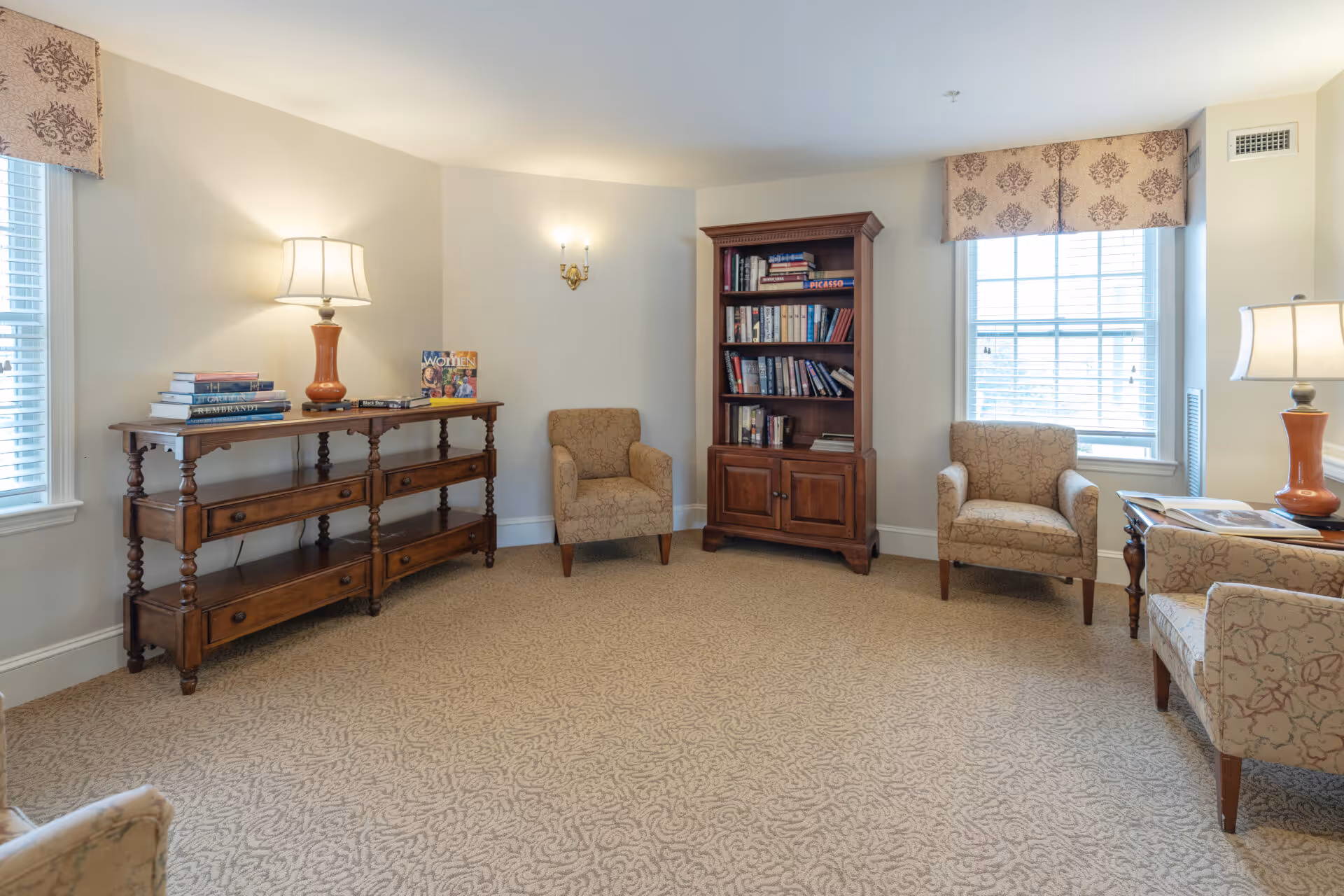 A cozy living room area with beige patterned carpet and light-colored walls. The room features two upholstered armchairs, a wooden bookshelf filled with books, a wooden console table with a lamp and books on it, and two windows with patterned valances allowing natural light to enter.