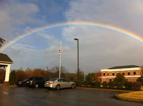 Rainbow arcs over a parking lot in front of a brick senior residence with flagpole.