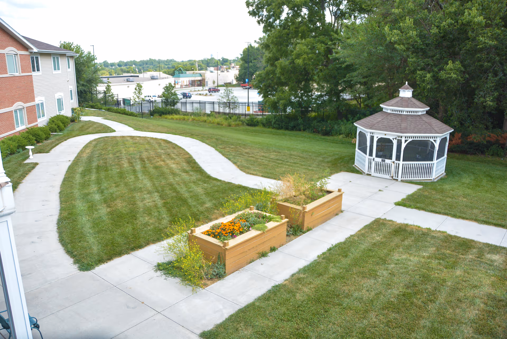 Outdoor view of a senior living facility showing a well-maintained grassy area with concrete walkways, two raised garden beds with flowers and plants, a white gazebo, and part of a multi-story building on the left side. Trees and a parking lot are visible in the background.