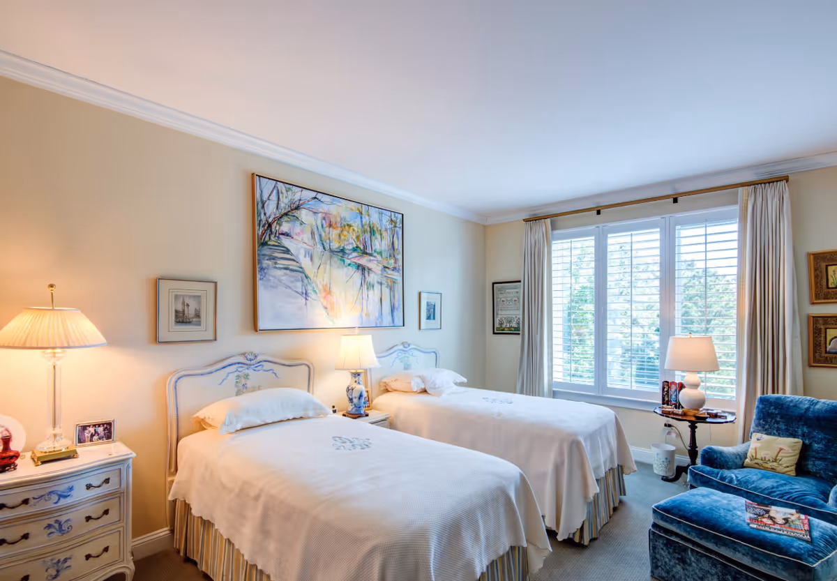 A bright and cozy bedroom with two twin beds covered in white bedspreads. Each bed has a decorative headboard and a pillow. Between the beds is a small nightstand with a blue and white lamp. The room features a large window with white shutters and beige curtains, allowing natural light to fill the space. There is a blue upholstered armchair with a matching ottoman near the window, and framed artwork adorns the walls.