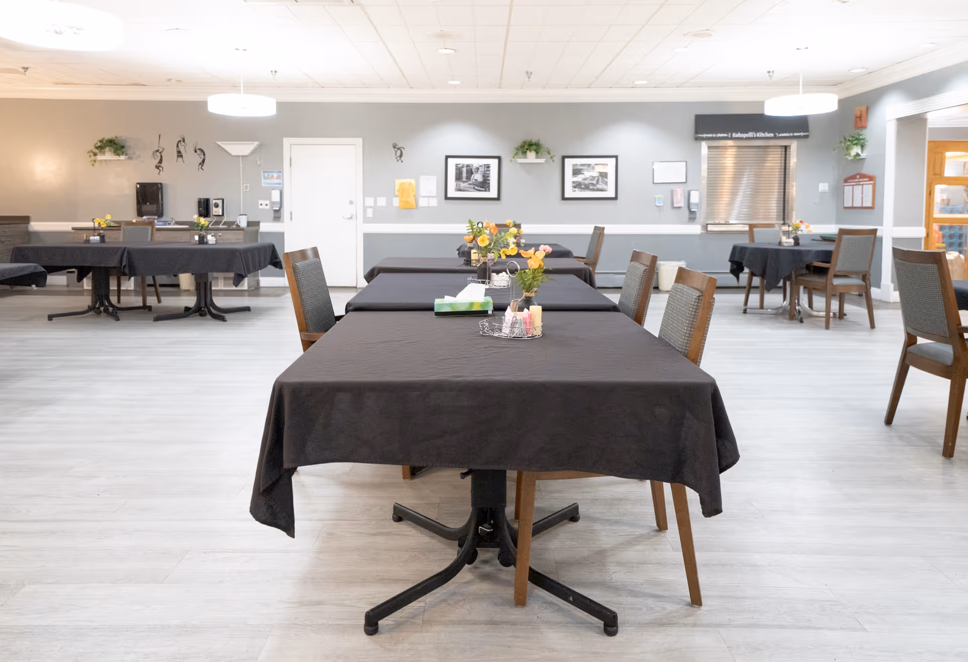 A spacious dining room with several tables covered in black tablecloths, each adorned with small flower arrangements and tissue boxes. The room has light-colored flooring and walls painted in neutral tones. There are chairs around the tables, framed pictures on the walls, and a serving window with a metal shutter. The room is well-lit with ceiling lights.