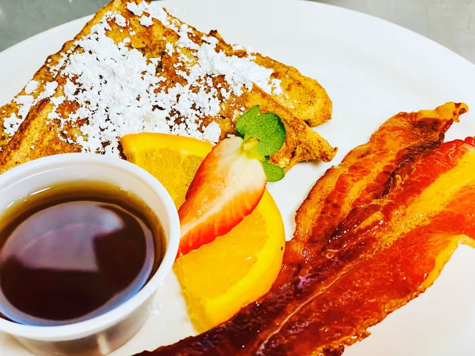 A breakfast plate with two strips of crispy bacon, a slice of French toast dusted with powdered sugar, a strawberry half, an orange slice, and a small cup of syrup.