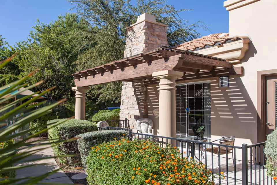 Outdoor patio area at Tuscany at McCormick Ranch featuring a stone fireplace with a wooden pergola overhead, cushioned chairs, a small table, and surrounded by green bushes and orange flowers under a clear blue sky.