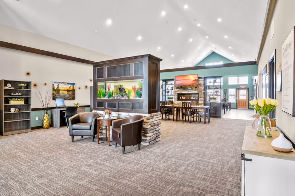 Spacious senior living facility common area with high vaulted ceilings and recessed lighting. The room features a large built-in aquarium divider with stone accents, two leather armchairs and a small round table in the foreground. In the background, there is a dining area with tables and chairs, a stone fireplace with a mounted TV above it, and large windows letting in natural light. To the left, there is a desk with a computer and a bookshelf with decorative items. On the right side, a console table holds a vase with yellow tulips and other decor items.