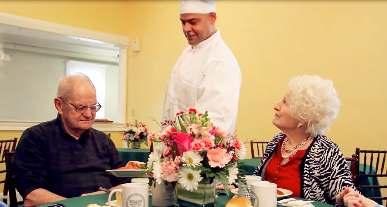 A chef in white uniform serves food to an elderly man and woman seated at a dining table with a green tablecloth and a floral centerpiece in a well-lit room.