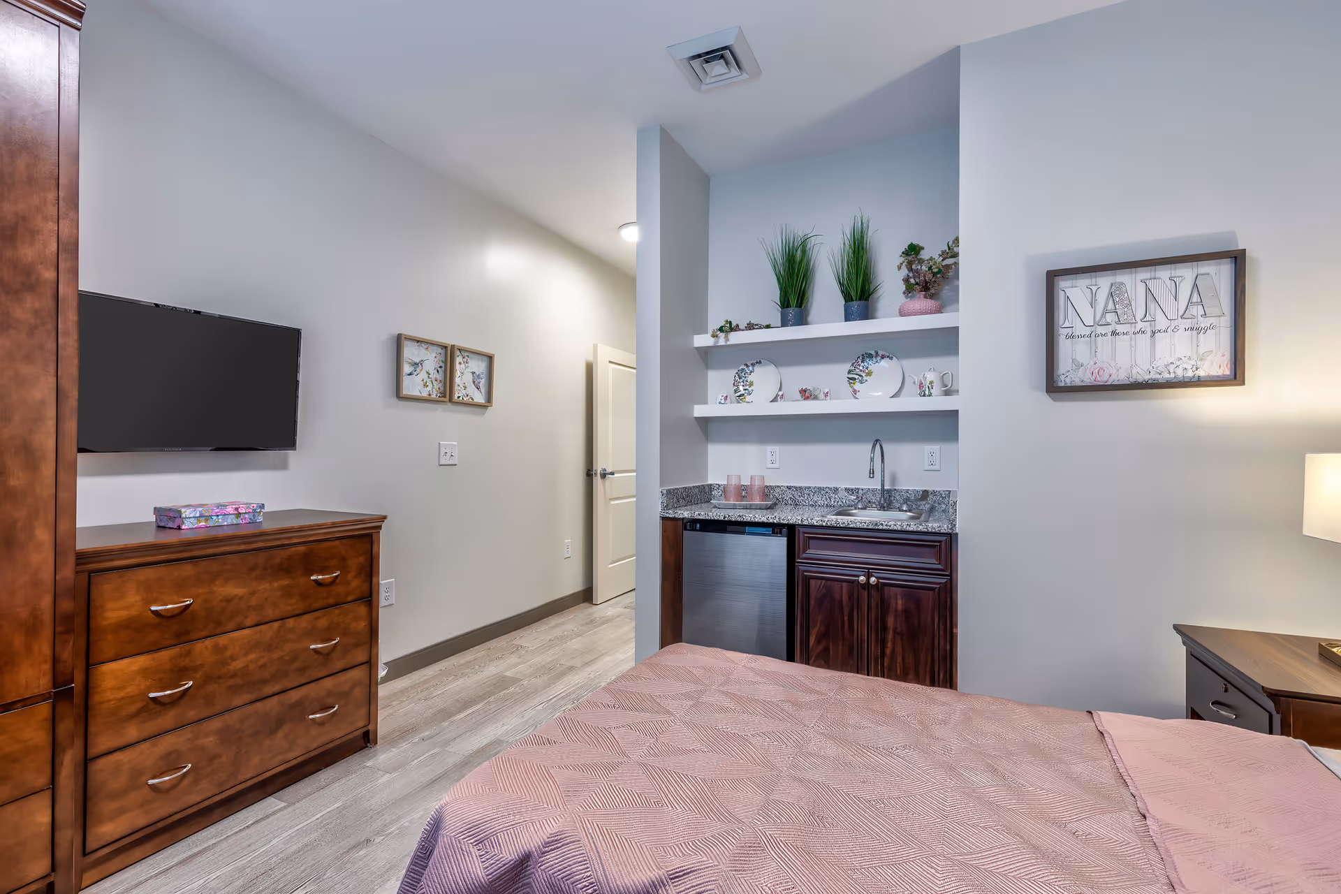 A cozy bedroom featuring a bed with a pink quilted bedspread, a wooden dresser with a flat-screen TV mounted above it, and a small kitchenette area with a sink, mini fridge, and open shelves decorated with plants and plates. The walls are light gray, and there is a framed sign on the wall that reads 'NANA'.