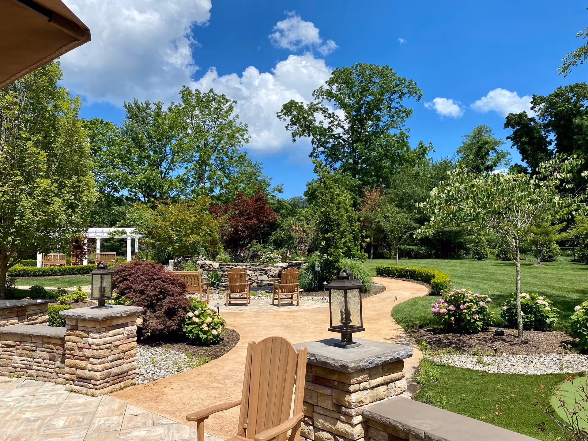 A beautifully landscaped outdoor garden area with a paved walkway, wooden chairs arranged around a small stone wall, various green trees and shrubs, and a bright blue sky with scattered white clouds.