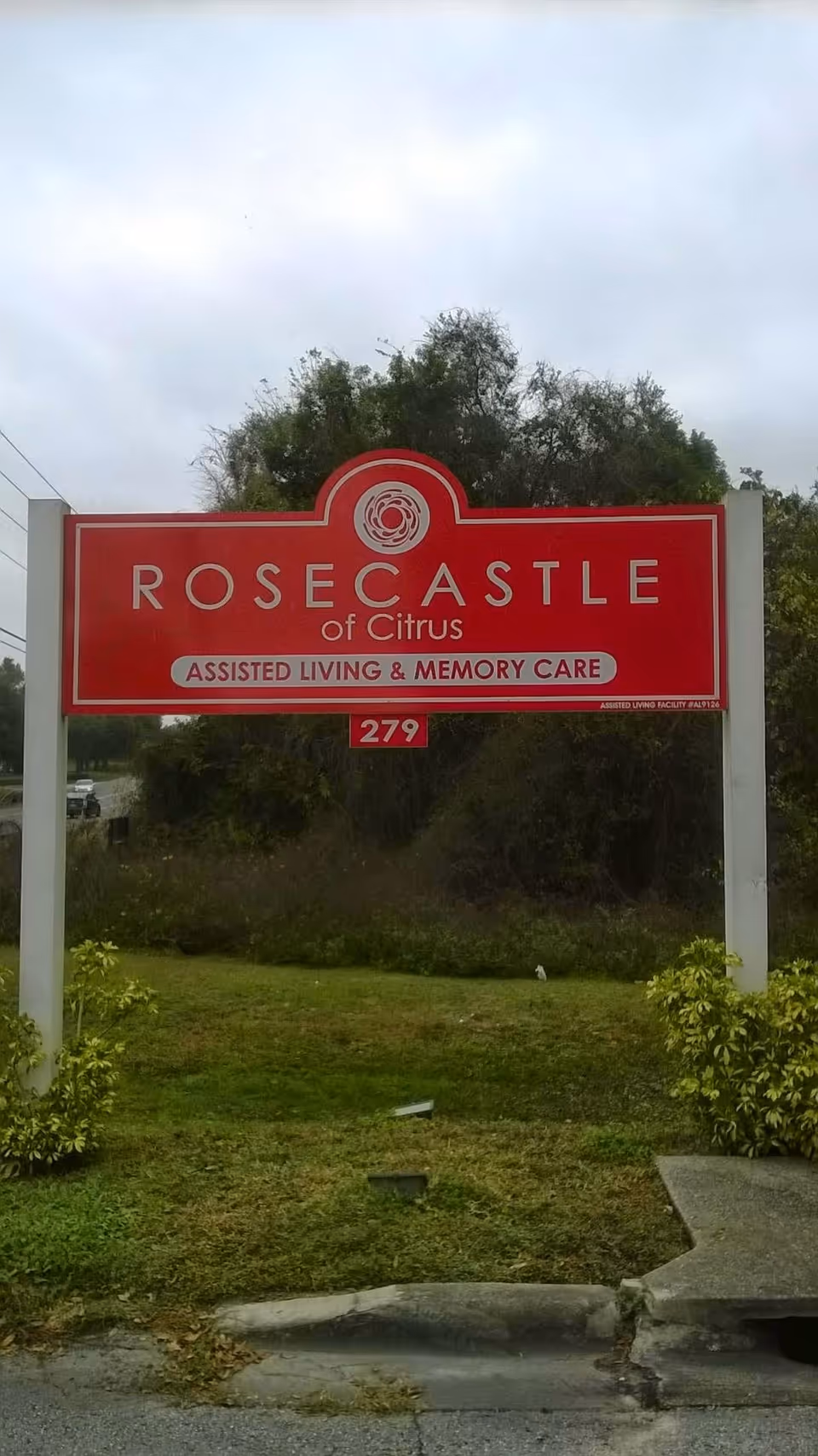A red and white outdoor sign for Rosecastle of Citrus, an assisted living and memory care facility, standing on a grassy area with bushes and trees in the background under a cloudy sky.