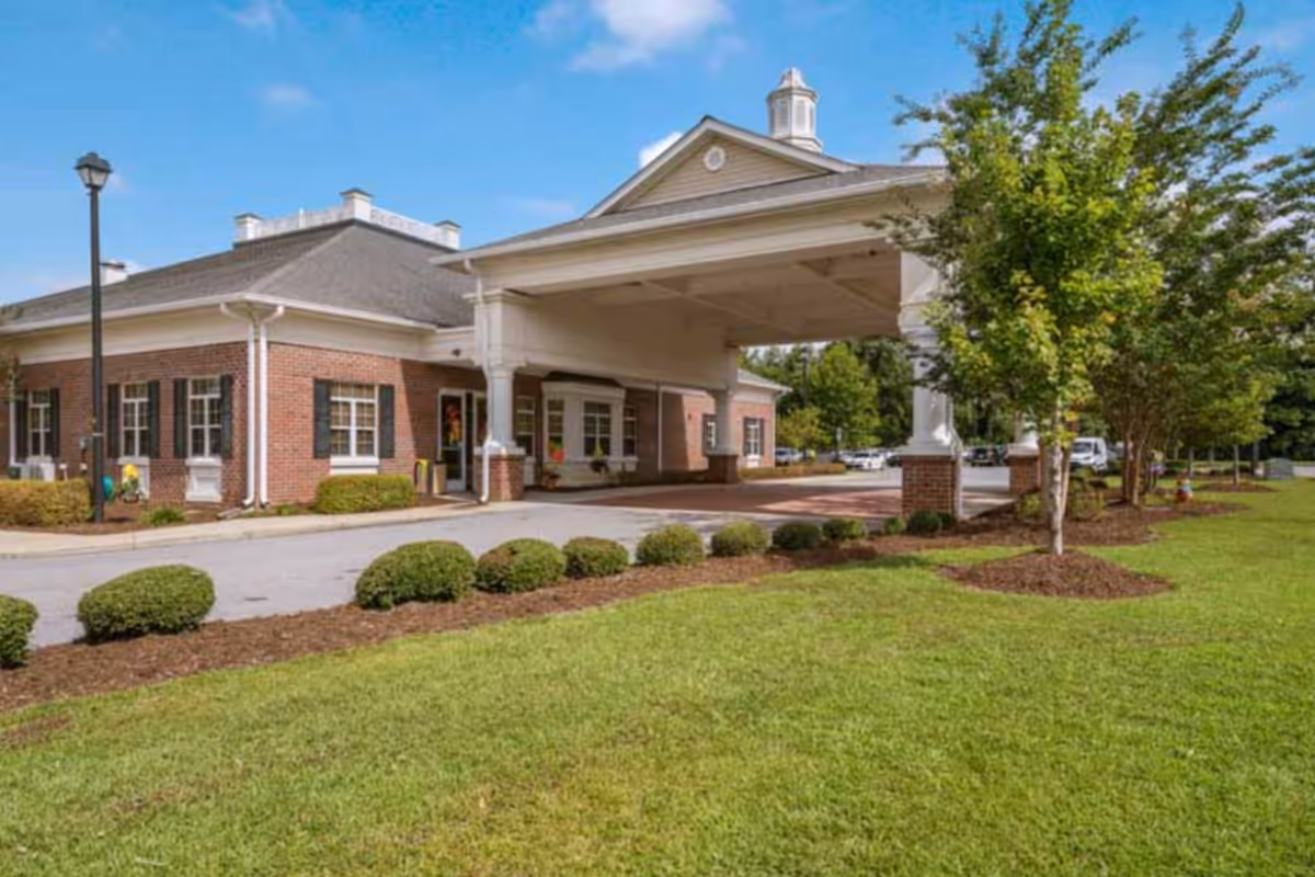 Exterior view of MacGregor Downs Health Center showing a brick building with white trim and a covered entrance driveway. The building is surrounded by neatly trimmed bushes, green grass, and trees under a clear blue sky.