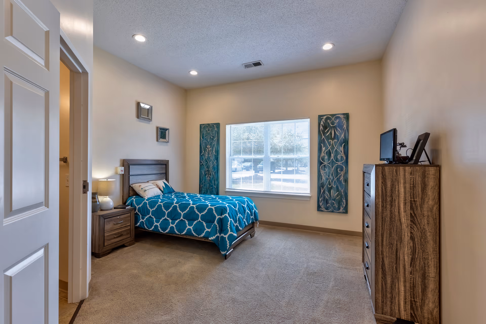 A bedroom with a single bed covered in a blue and white patterned comforter. There is a wooden nightstand with a lamp next to the bed, and a wooden dresser with a small TV on top. The room has beige walls, a large window with blinds, and two decorative wall panels on either side of the window. The door to an adjacent bathroom is open, showing a glimpse of the bathroom interior.