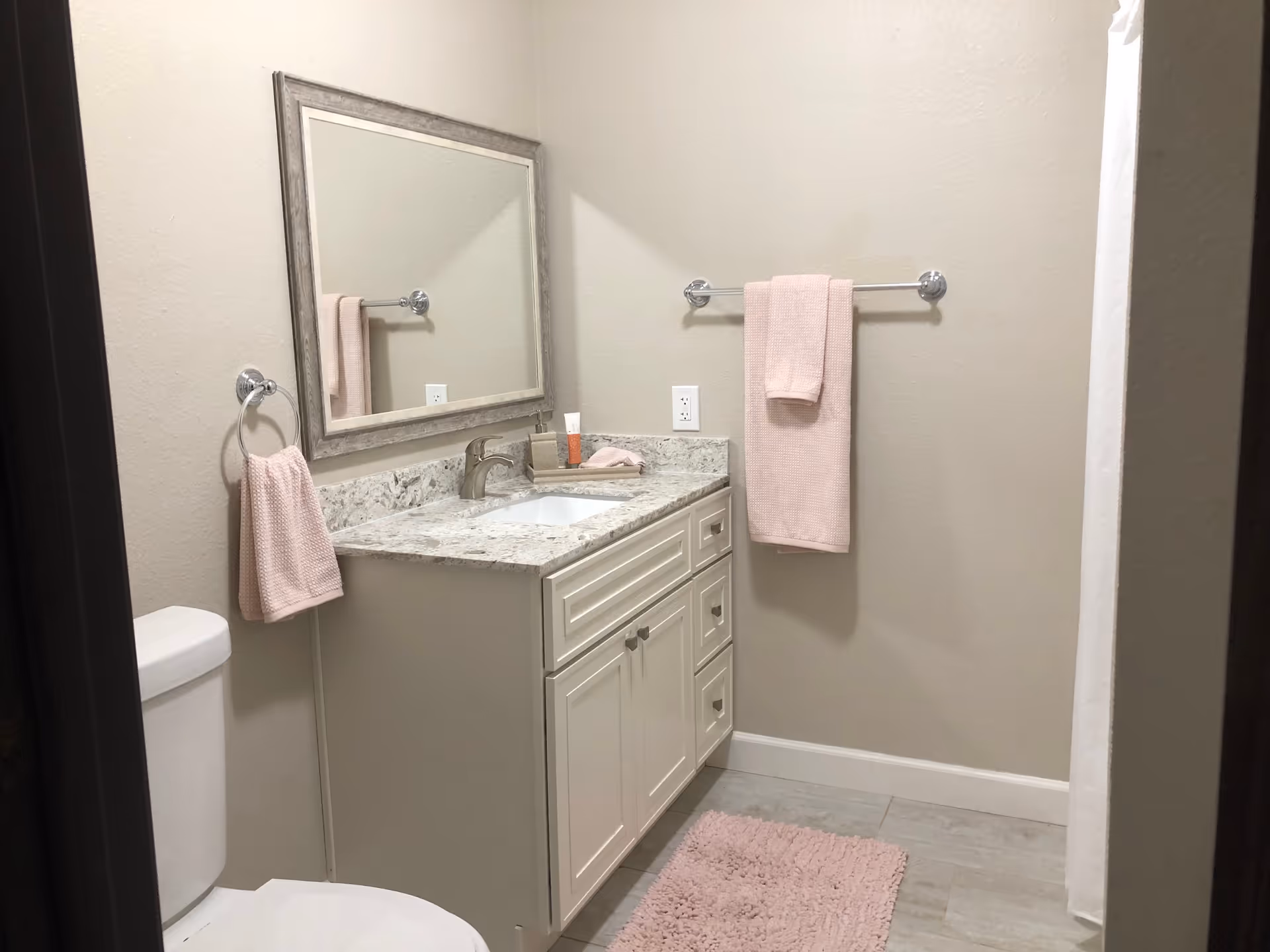 A clean bathroom with a white toilet, a white vanity with a granite countertop and a sink, a large rectangular mirror above the vanity, pink towels hanging on a towel ring and towel bar, a pink bath mat on the floor, and a white shower curtain on the right side.
