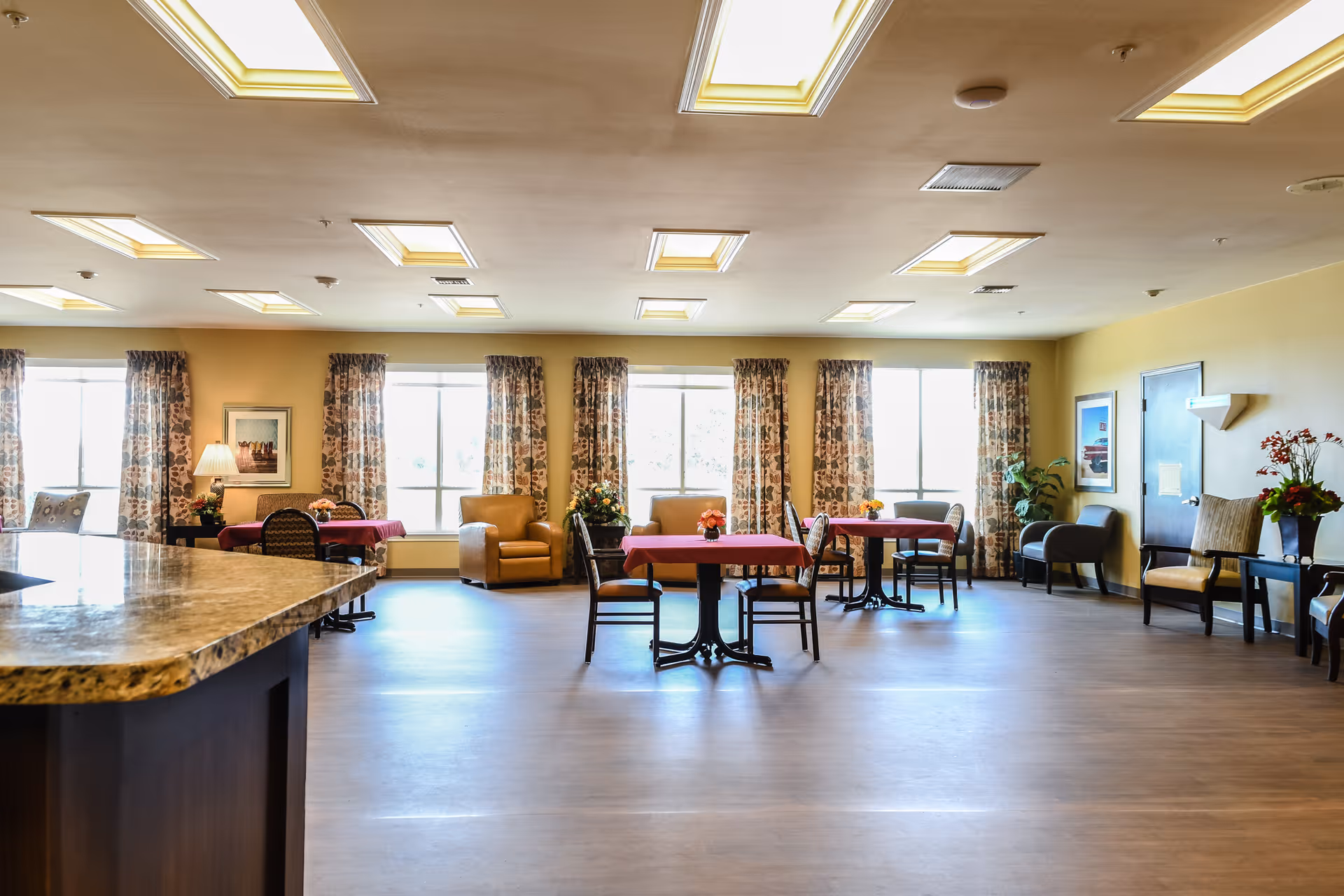 A spacious and well-lit common area with multiple tables covered in red tablecloths, surrounded by chairs. The room has large windows with floral curtains letting in natural light. There are several armchairs and side tables with lamps and flower arrangements along the walls. The ceiling has recessed lighting panels, and the floor is a light wood finish.