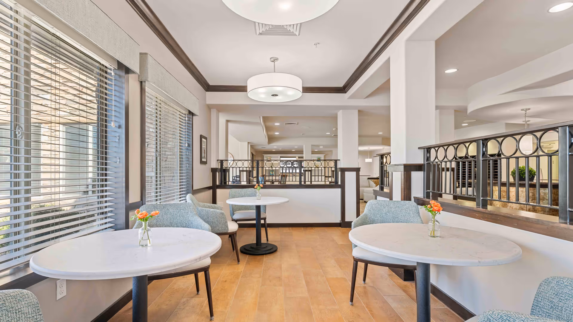A bright and airy common area with round white tables and light blue upholstered chairs. Each table has a small vase with orange flowers. Large windows with blinds allow natural light to fill the space. The room features light wood flooring, white walls with dark trim, and modern ceiling lights. A decorative black metal railing separates this seating area from another part of the room.