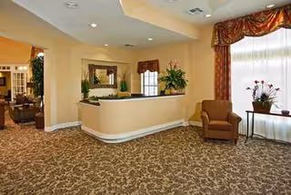 Senior living facility lobby with curved reception desk, patterned carpet, plants, and seating.