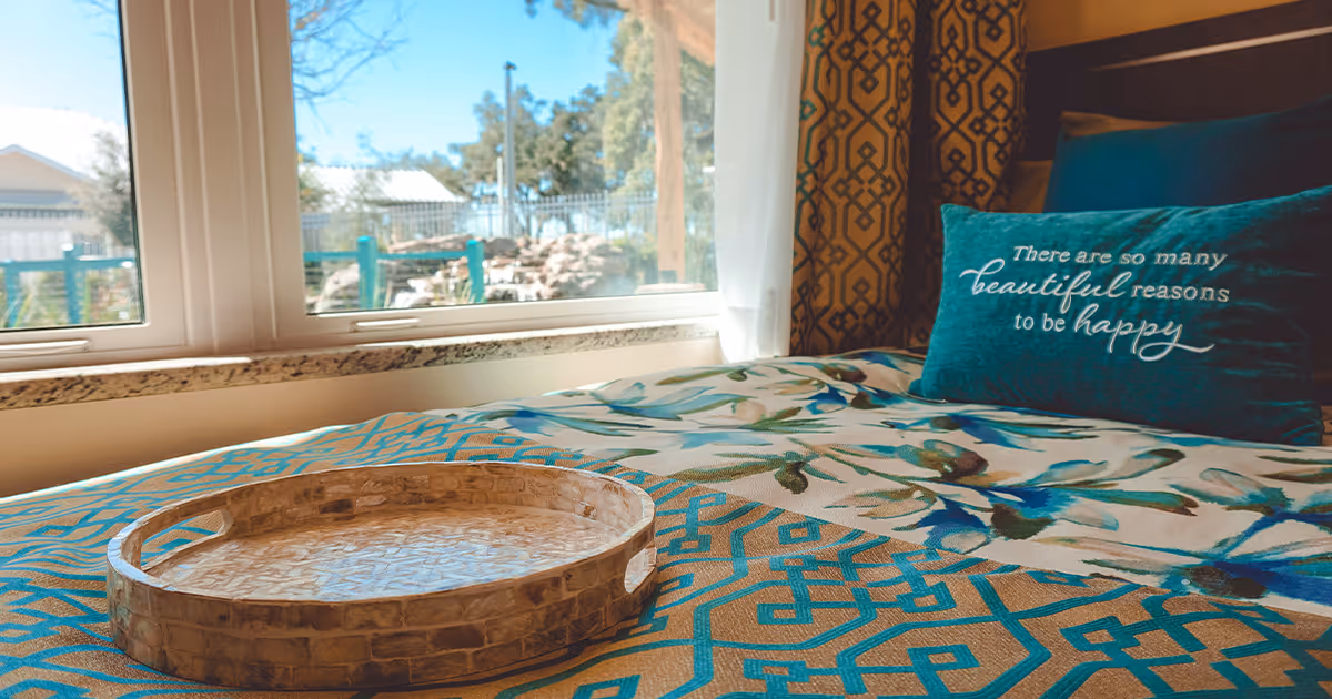 Sunlit bedroom with a patterned bedspread, a decorative round tray on the bed, and a teal pillow by the window that reads 'There are so many beautiful reasons to be happy'.
