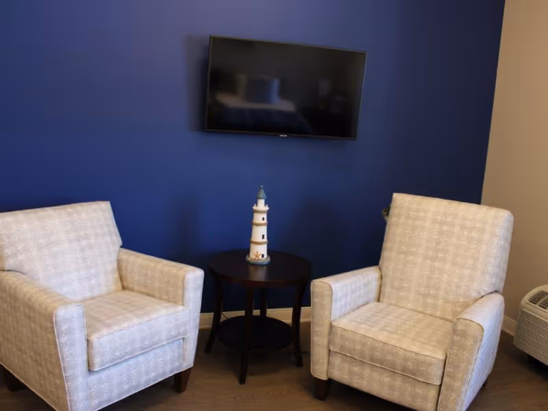 Two beige upholstered armchairs positioned on either side of a small round dark wooden table with a decorative lighthouse figurine on top, set against a blue wall with a flat-screen TV mounted above.