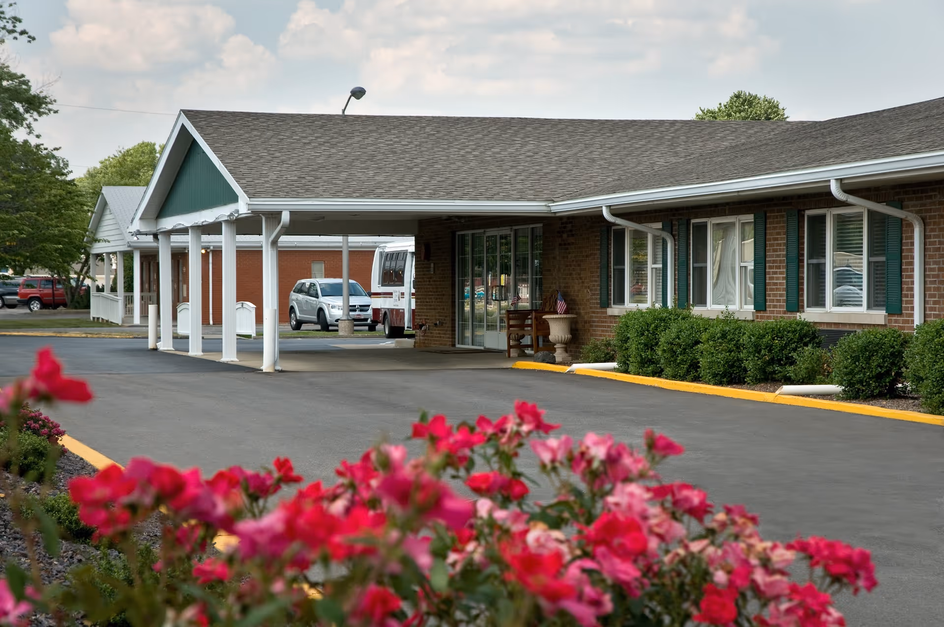 Exterior of a single-story brick senior living facility with a covered entrance drive and pink flowers in the foreground.