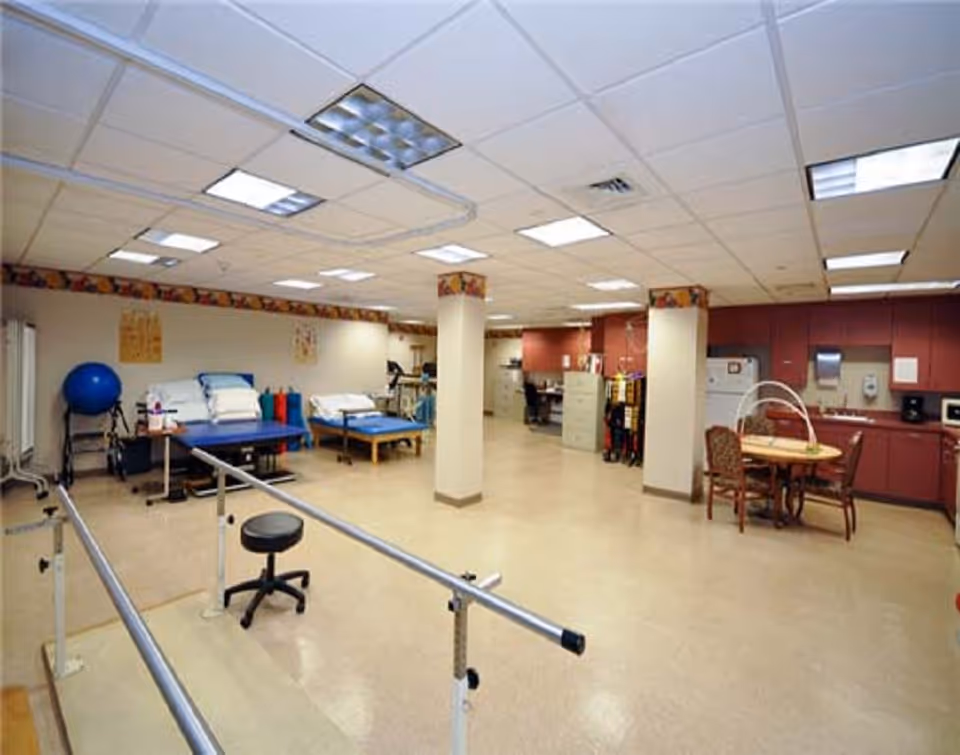 A spacious rehabilitation room with parallel bars for walking exercises, a stool, two therapy beds with pillows, exercise equipment including a large blue exercise ball, and a kitchenette area with a table and chairs. The room has a tiled ceiling with fluorescent lighting and beige flooring.