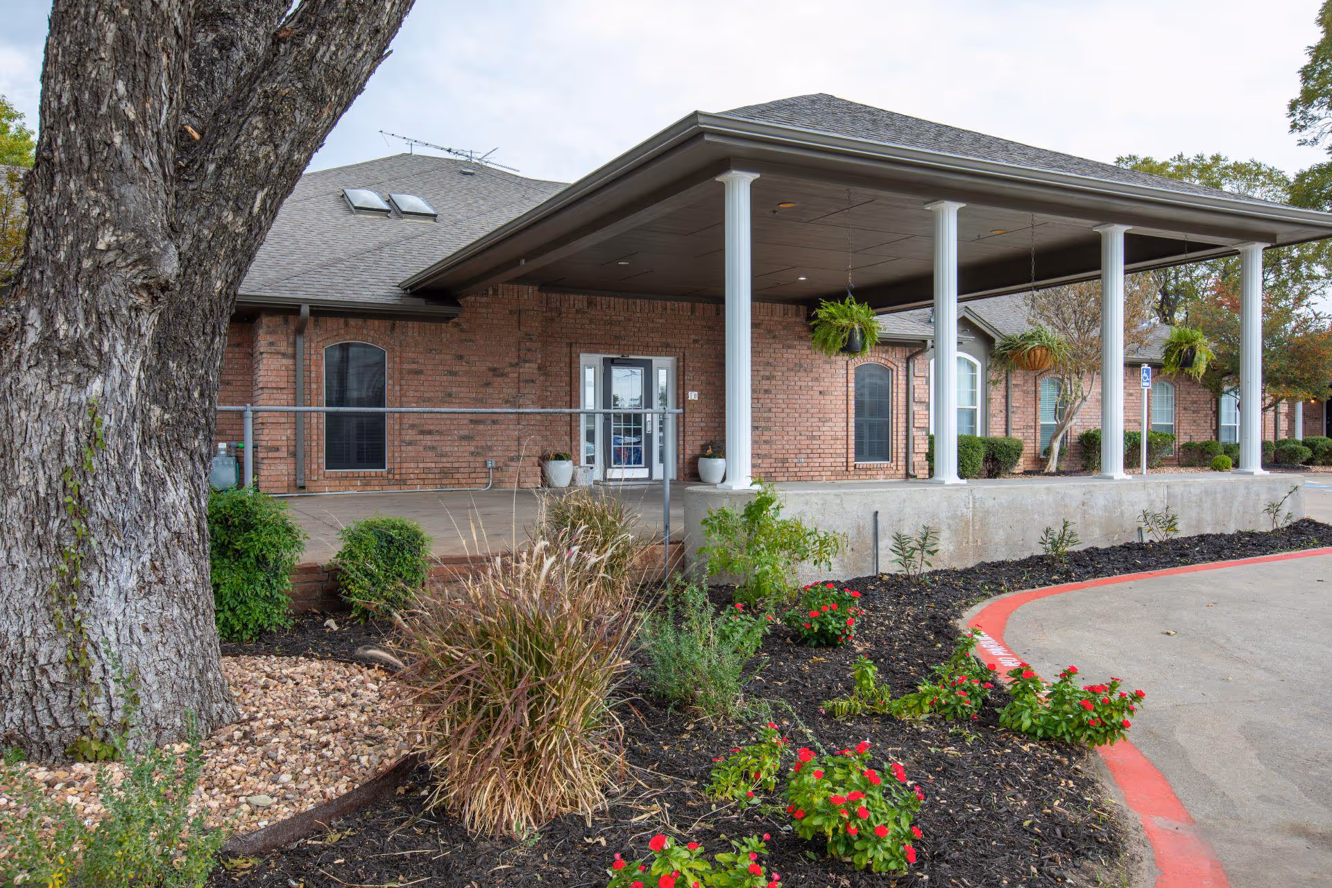 Exterior view of a brick building with a covered entrance supported by white columns. There are hanging plants under the roof of the entrance and landscaped flower beds with red flowers and shrubs in front. A large tree trunk is visible on the left side of the image.