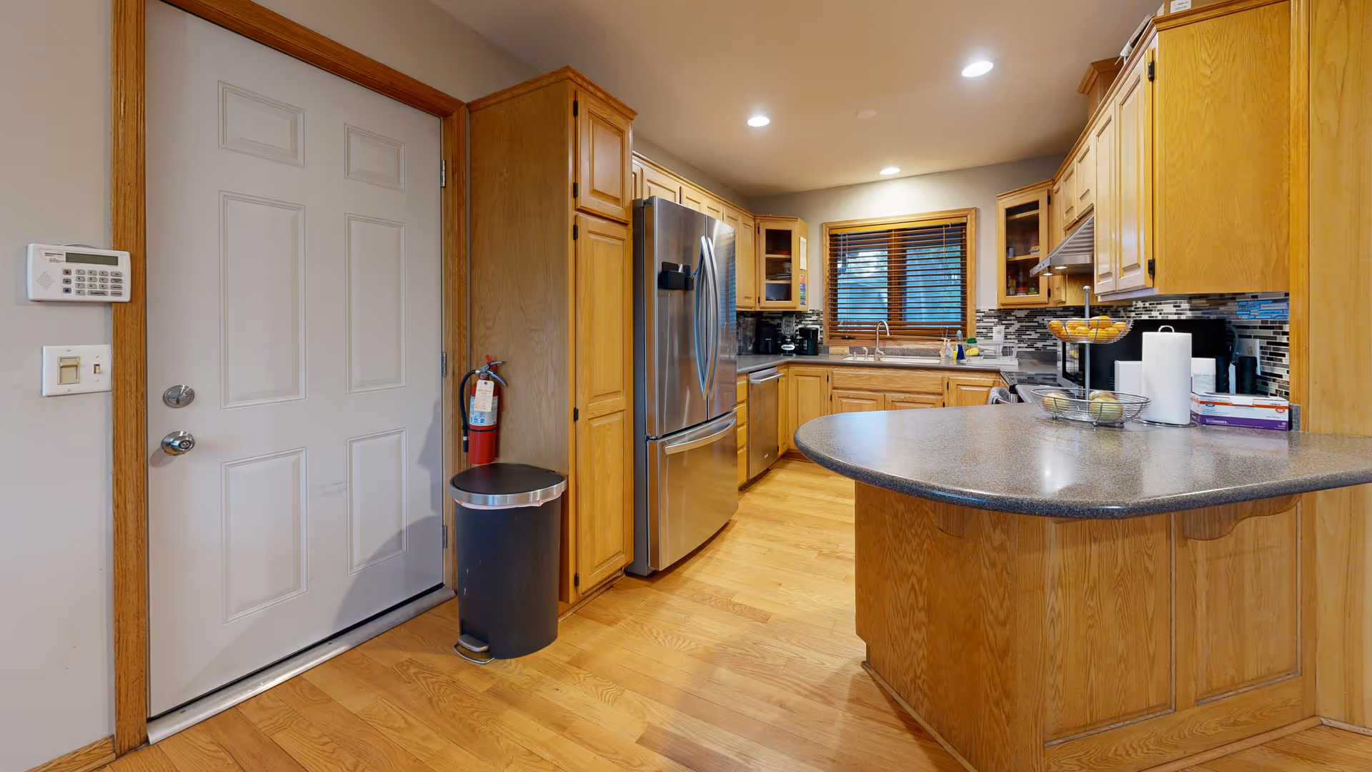 A modern kitchen with wooden cabinets and hardwood floors. The kitchen features a stainless steel refrigerator, a dishwasher, a sink under a window with wooden blinds, and a curved countertop island with various items including a paper towel holder and fruit baskets. A white door with a keypad and a fire extinguisher next to a trash can are visible on the left side.