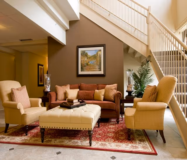 A cozy sitting area with a brown sofa, two beige armchairs, and a large tufted ottoman on a red patterned rug. The space is under a staircase with white railings, and there is a framed painting on the brown accent wall behind the sofa. Two side tables with decorative lamps and a potted plant are also visible.