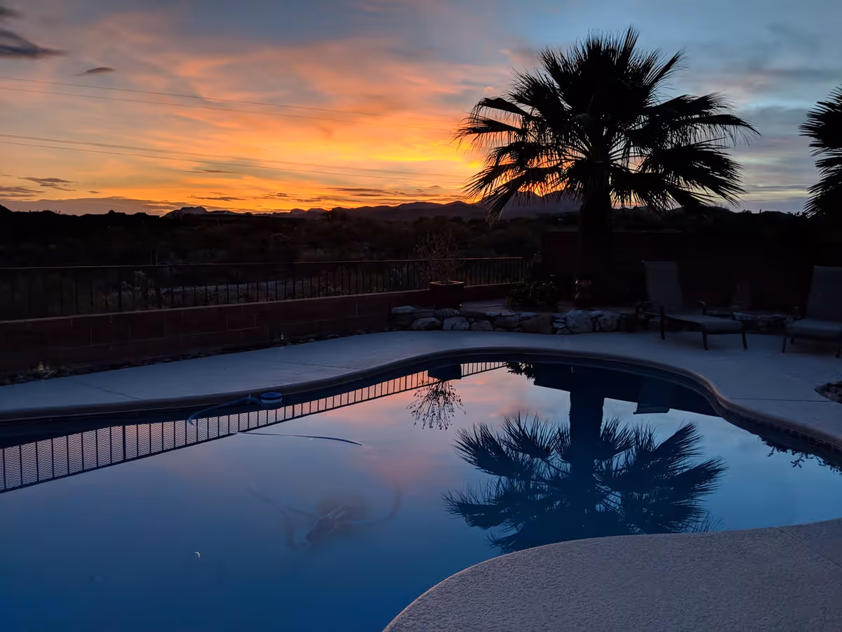 A swimming pool at sunset with a palm tree and lounge chairs nearby. The sky is colorful with shades of orange, yellow, and blue, and the palm tree is reflected in the calm water of the pool.