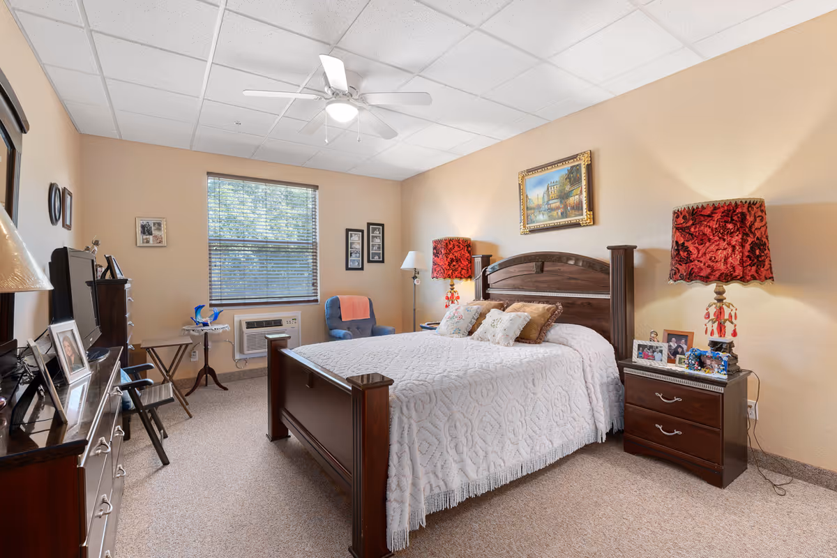 A cozy bedroom in an assisted living facility featuring a large wooden bed with a white quilt and decorative pillows. There are two dark wood nightstands with red patterned lamps on each side of the bed. A framed painting hangs above the bed. The room has beige walls, a window with blinds, a blue armchair with a coral throw, a ceiling fan with a light, and various framed photos on the furniture.