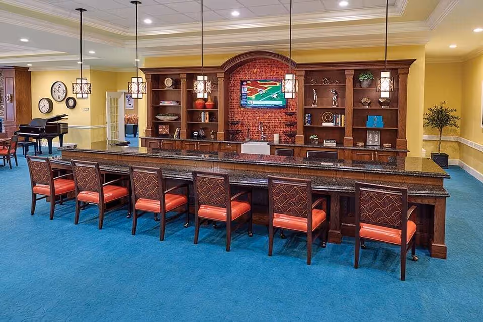 A spacious common area with a long dark granite countertop bar and eight wooden chairs with red cushions. Behind the bar is a wooden shelving unit with decorative items and a flat-screen TV mounted on a red brick accent wall. The room has blue carpet, yellow walls, and pendant lights hanging above the bar. In the background, there is a black grand piano and a wall clock.