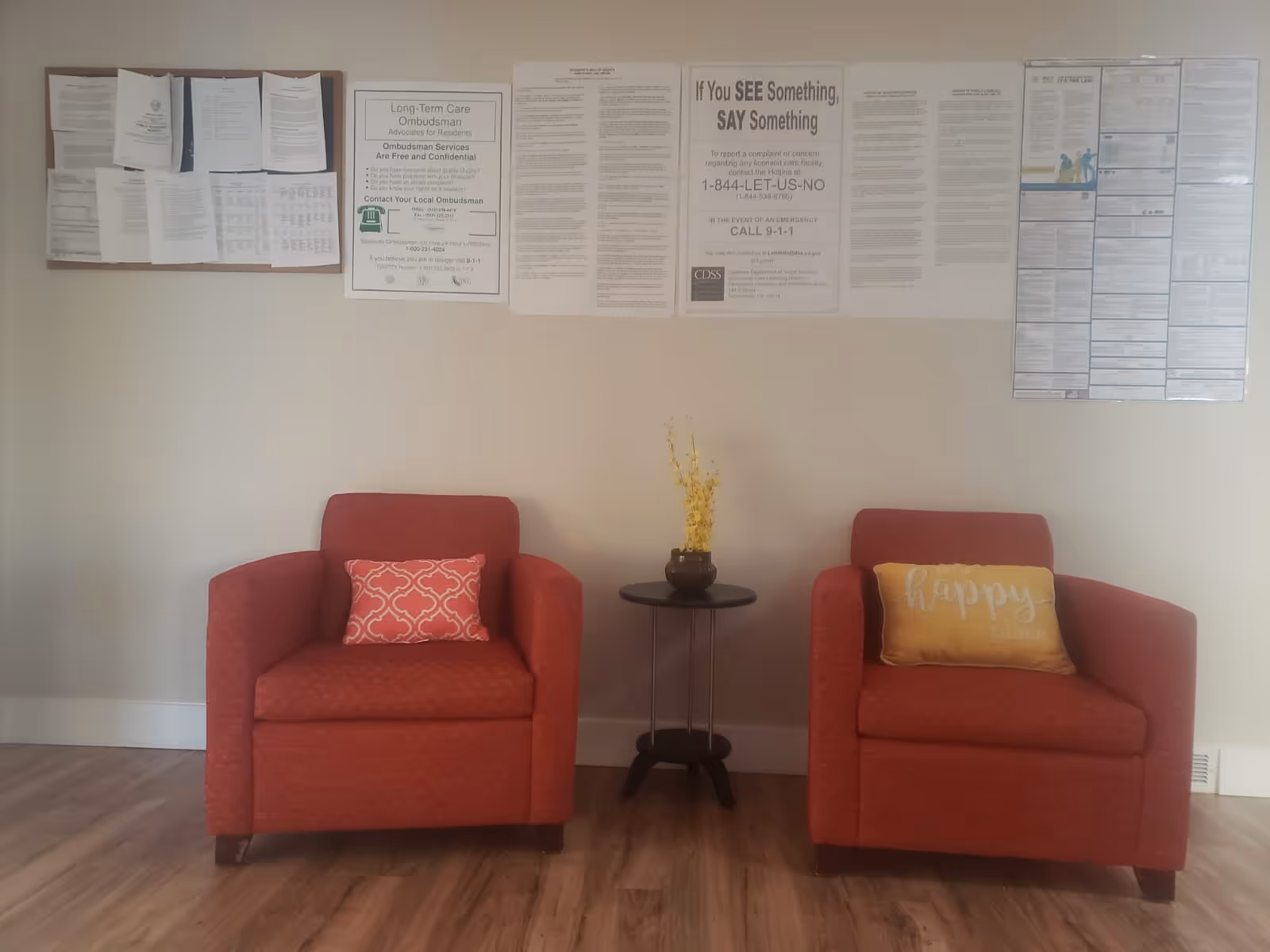 Two red armchairs with decorative pillows, one pink and one yellow with the word 'happy', are placed on either side of a small round table with a vase of yellow flowers. Behind the chairs, a wall displays several notices and informational posters. The floor is wooden.
