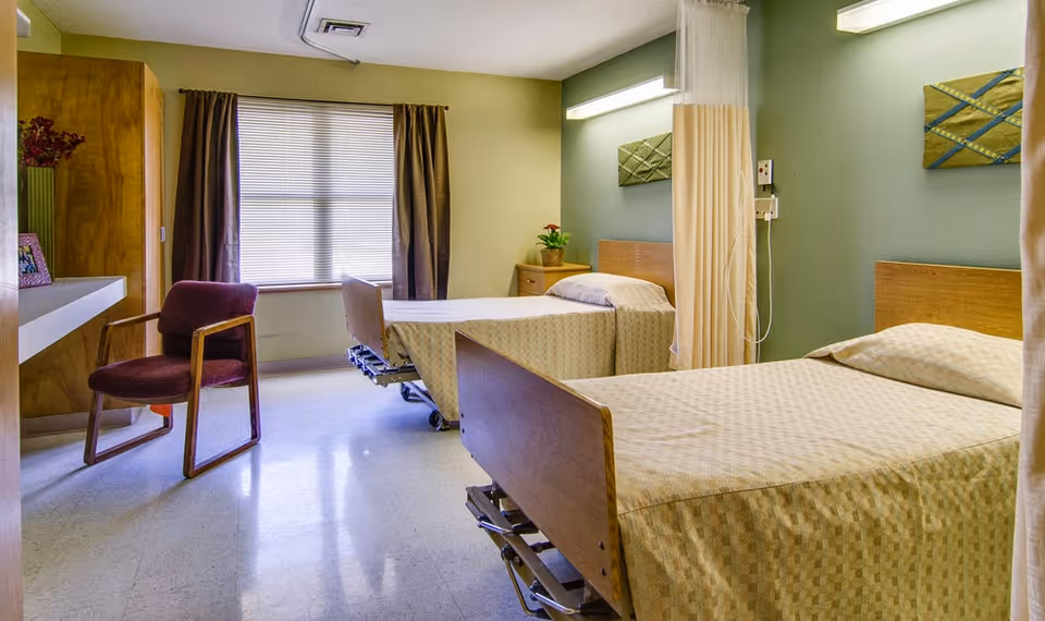 A room with two hospital-style beds with beige checkered bedspreads, a wooden chair with a purple cushion, a window with brown curtains, a small wooden nightstand with a potted plant, and a privacy curtain partially visible on the right side.