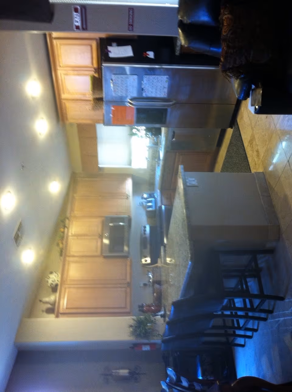 Large kitchen area with a central island, bar stools, stainless steel refrigerator, wooden cabinets and recessed ceiling lights.
