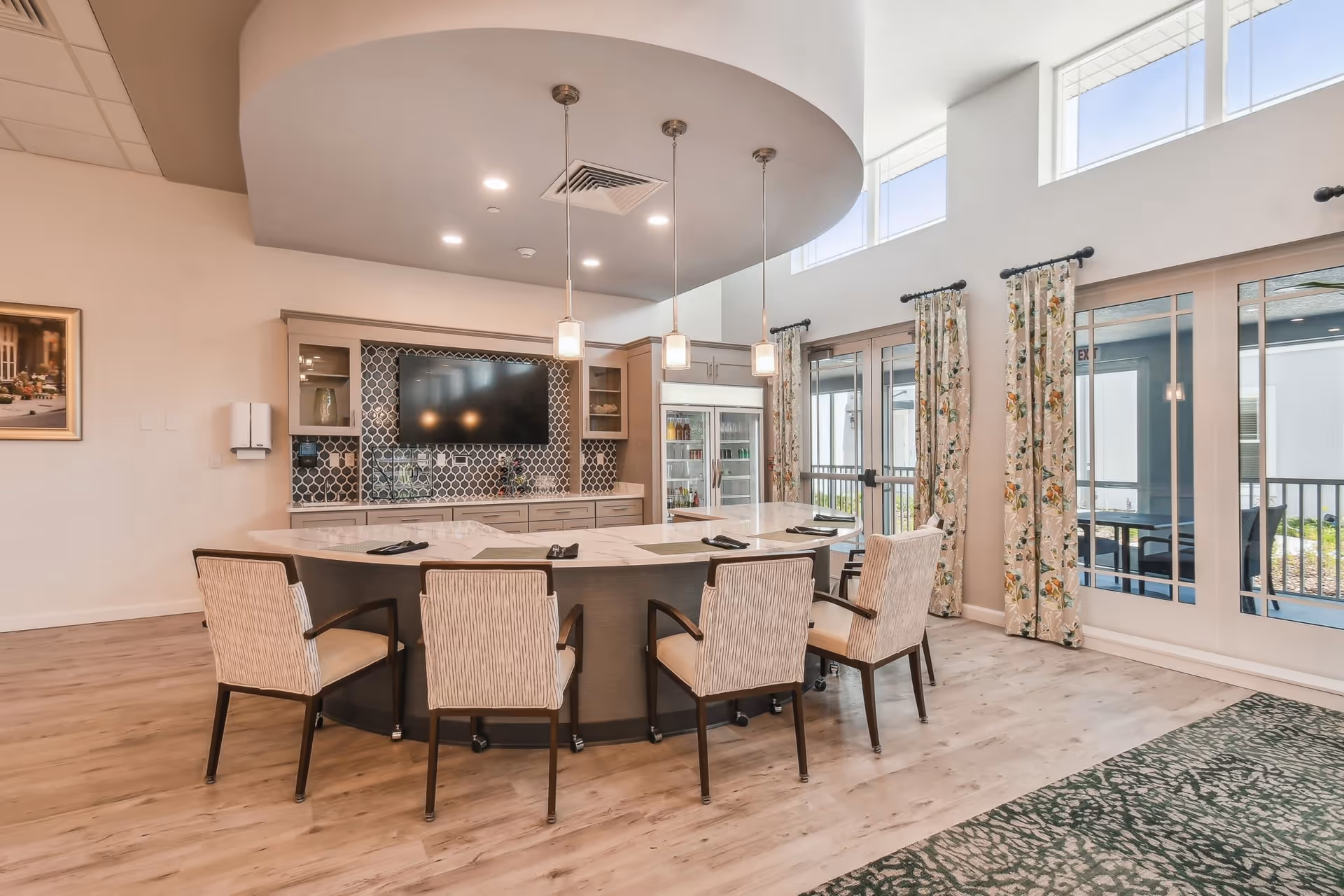A modern common area with a large curved marble countertop island surrounded by six cushioned chairs. Above the island hang three pendant lights. Behind the island is a wall with a large flat-screen TV mounted above a patterned tile backsplash and cabinetry. To the right, large windows and glass doors with floral curtains let in natural light and provide a view of an outdoor patio area.