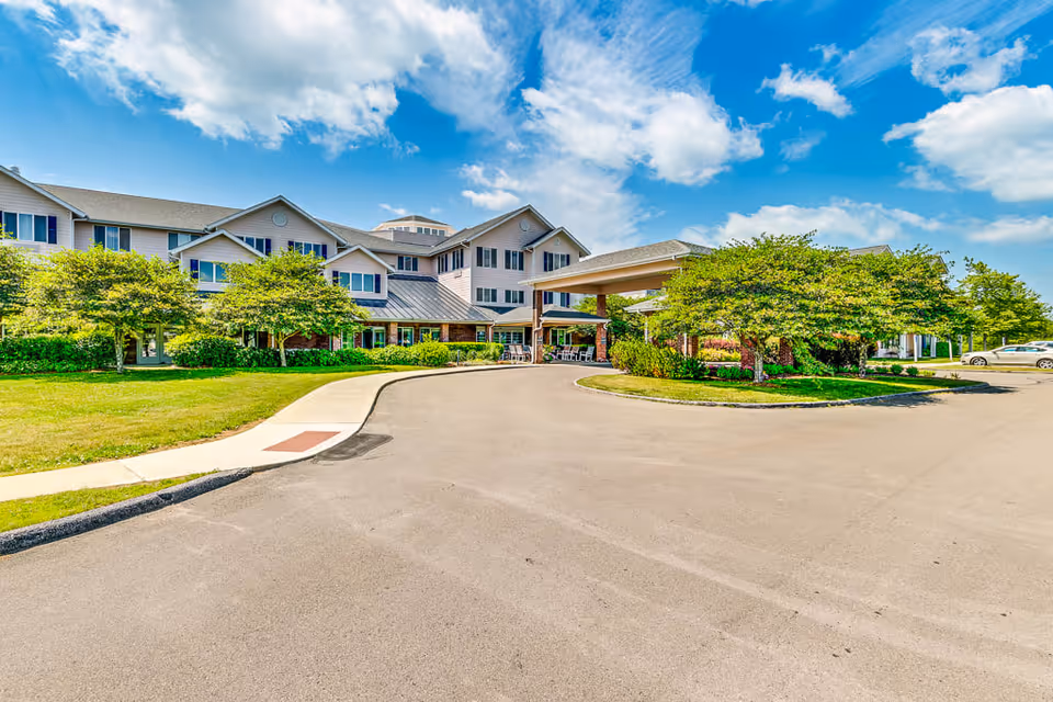 Exterior view of Solstice Senior Living at Bangor, showing a large multi-story building with a covered entrance driveway, surrounded by green trees and a well-maintained lawn under a partly cloudy blue sky.