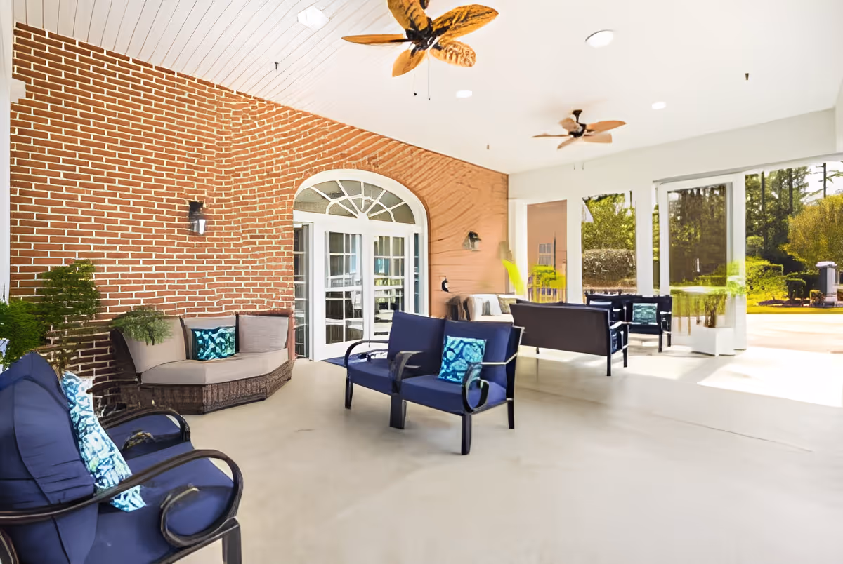 A bright and spacious covered patio area at Southern Pines Senior Living featuring comfortable seating with blue cushions and decorative pillows, a brick wall with a white arched door, ceiling fans with wooden blades, and large glass doors opening to a sunny outdoor garden space.