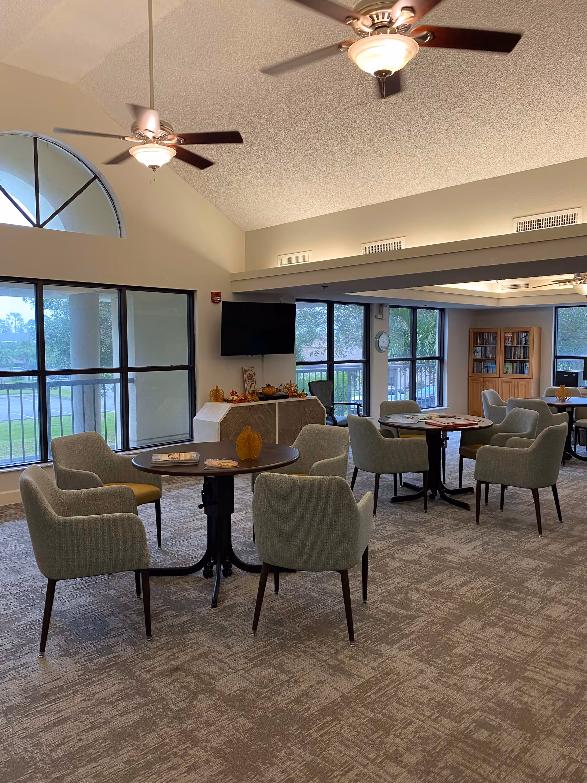 A spacious common area with multiple round tables surrounded by gray upholstered chairs. The room has large windows letting in natural light, a wall-mounted TV, ceiling fans with lights, and a bookshelf in the corner. The decor includes small autumn-themed decorations on a cabinet beneath the TV.