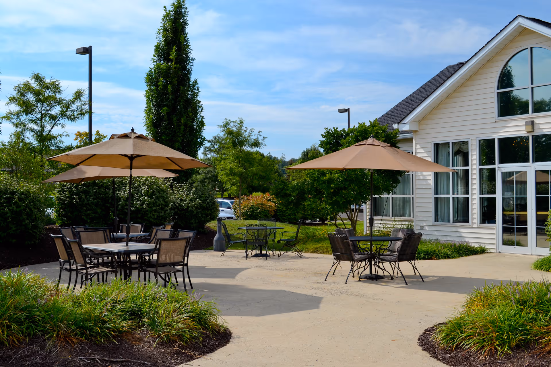 Outdoor patio area with several tables and chairs under large beige umbrellas, surrounded by greenery and bushes, next to a building with large windows and a glass door.