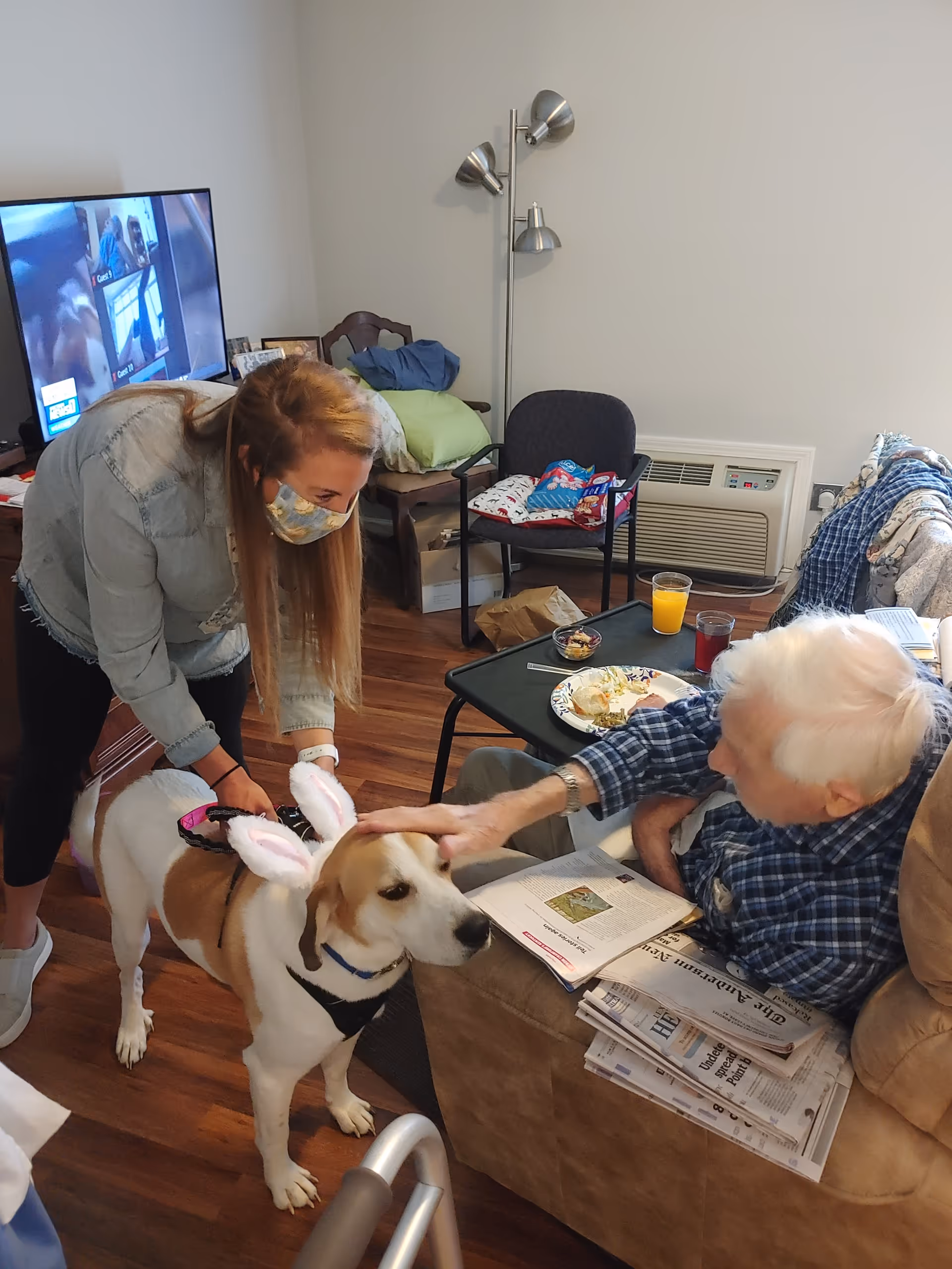 An elderly man sitting in a recliner chair inside a living room area, petting a dog wearing bunny ears. A woman wearing a face mask is holding the dog’s leash and smiling. The room has wooden floors, a TV in the background, a small table with food and drinks, and some chairs with cushions.