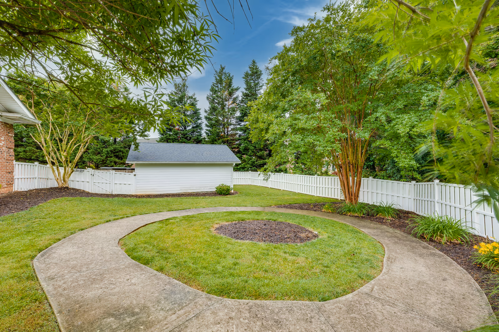 A circular concrete pathway surrounds a grassy area with a small patch of mulch in the center. The scene is outdoors with green trees and shrubs along a white wooden fence. A small white shed with a gray roof is visible in the background under a partly cloudy blue sky.