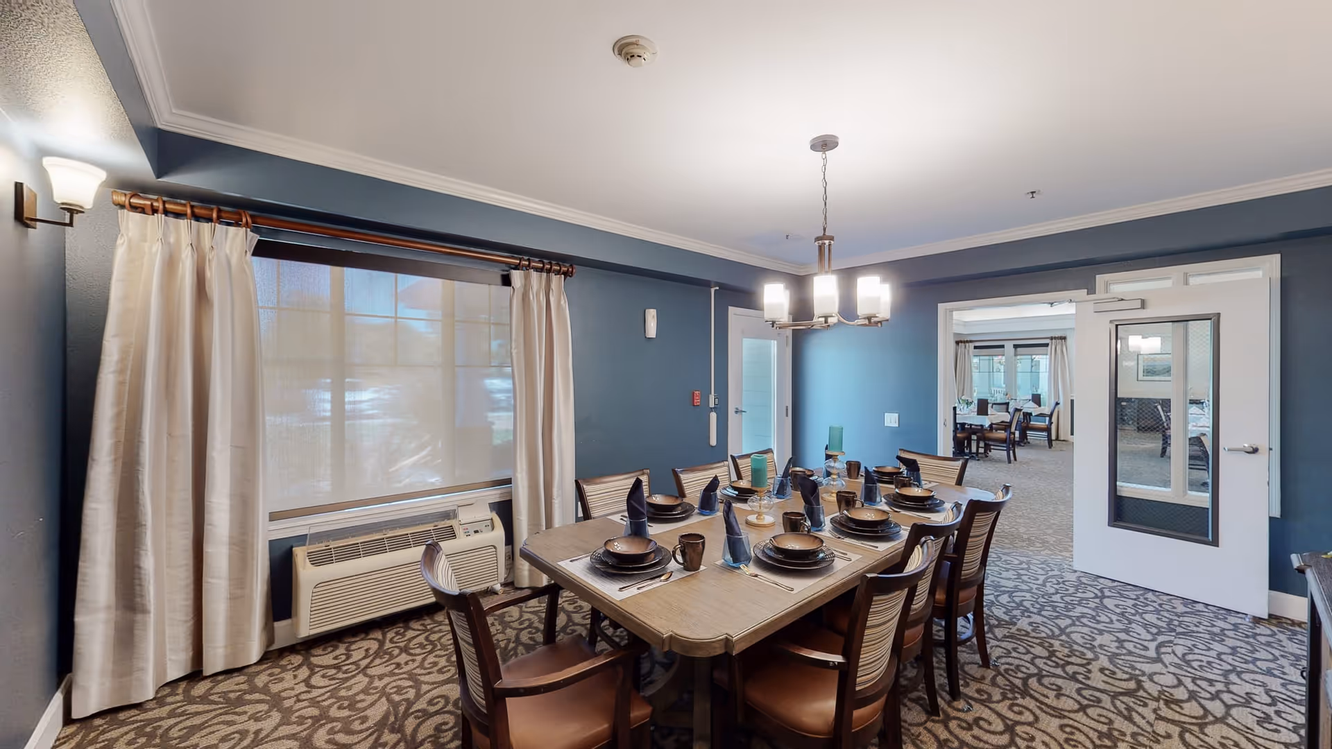 A dining room with a rectangular wooden table set for eight people with plates, bowls, mugs, and folded napkins. The room has blue walls, a patterned carpet, a large window with white curtains, and a modern chandelier hanging above the table. There is a doorway leading to another room with more dining tables and chairs.