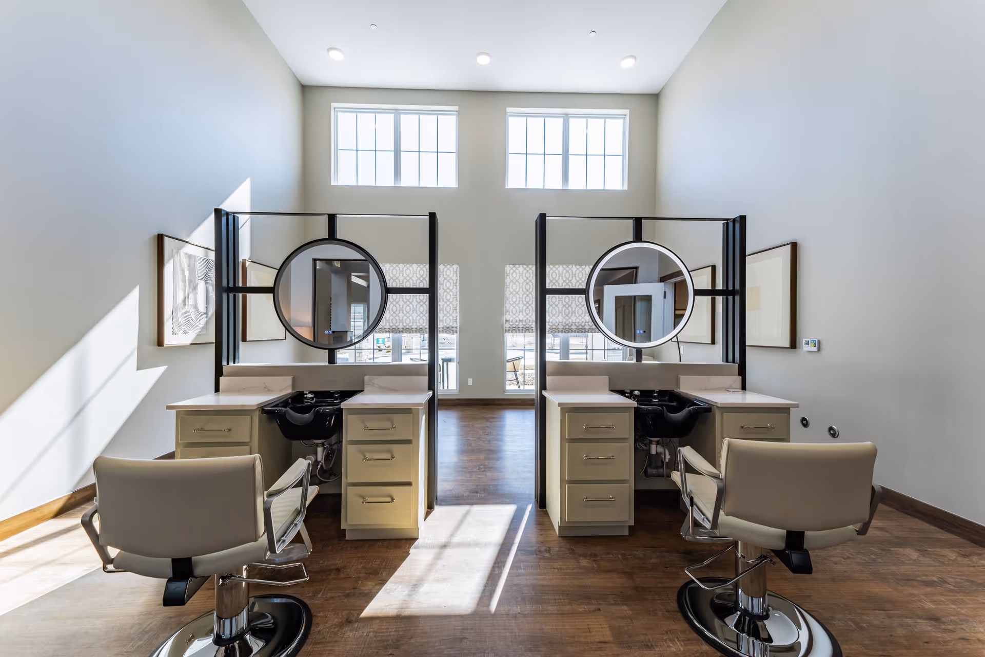 Interior view of a senior living facility hair salon area with two styling stations. Each station has a round mirror, a black sink, beige cabinetry with drawers, and a beige salon chair. Large windows let in natural light, and the walls are painted light gray with framed artwork on either side.