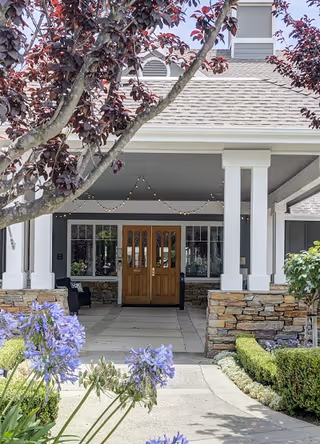 Entrance of a building with double wooden doors under a covered porch supported by white columns. There are stone walls on either side of the walkway leading to the doors, with purple flowers and green shrubs in the foreground. A tree with dark red leaves partially frames the top of the image.