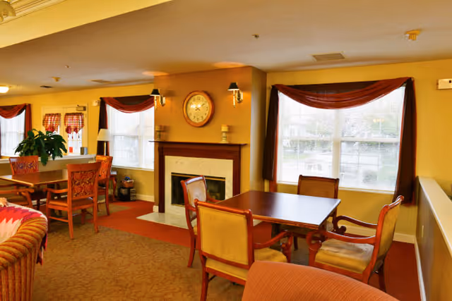 A cozy living room area with a fireplace, a clock above it, and several wooden tables and chairs arranged near large windows with red curtains. The room has warm yellow walls and carpeted floors, creating a welcoming atmosphere.