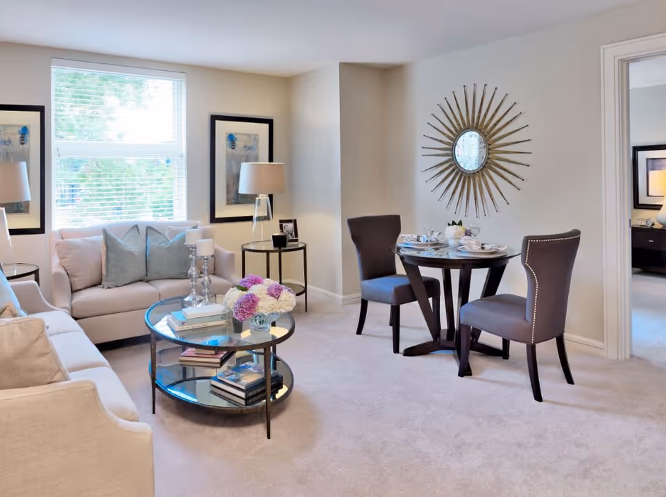 A bright and cozy living room with two beige sofas adorned with light blue and beige pillows. A round glass coffee table with books and a vase of flowers sits in the center. Two side tables with lamps flank the sofas. In the corner, there is a small round dining table set for two with dark upholstered chairs. A decorative sunburst mirror hangs on the wall above the dining table. The room has light-colored walls and carpeted flooring, with a window letting in natural light.